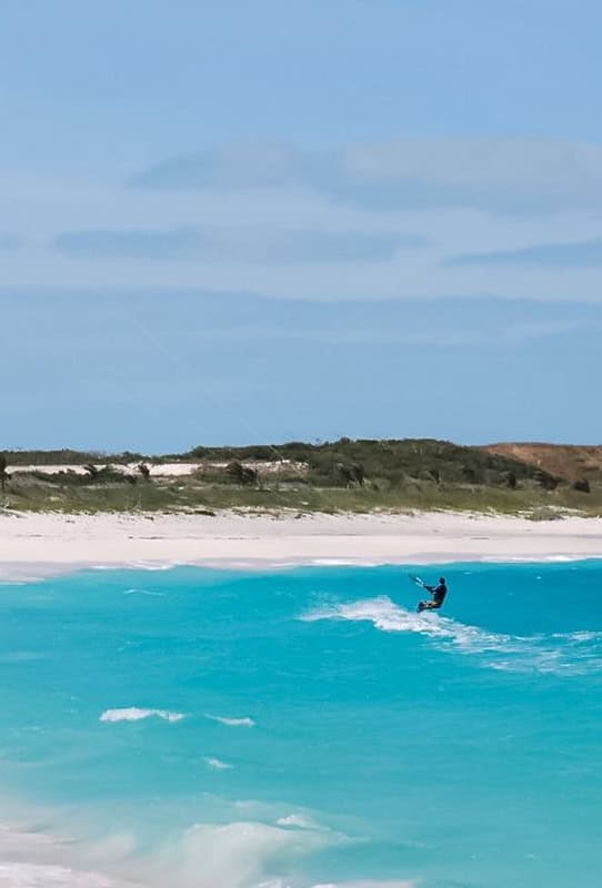 Kiteboarder riding a turquoise wave near a white-sand beach with low dunes under a blue sky.