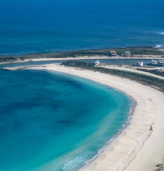 Aerial view of a crescent-shaped white-sand beach with vivid turquoise water and a small road with houses along the coast.